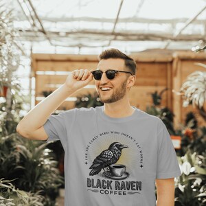 May include: A gray t-shirt with a black raven illustration holding a cup of coffee. The text on the shirt reads "For the early bird who doesn't eat worms. Black Raven Coffee."