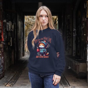 May include: A navy blue sweatshirt with a cartoon cat wearing a hoodie and holding a cup of coffee. The text on the sweatshirt reads "Here for the Nine Lives and Coffee Breaks".