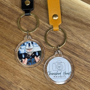 May include: Two keychains with clear acrylic charms. One keychain has a black leather strap and a photo of a football player. The other has a yellow leather strap and a charm with a camera graphic and the text "Cherished Heart MAGNETS".
