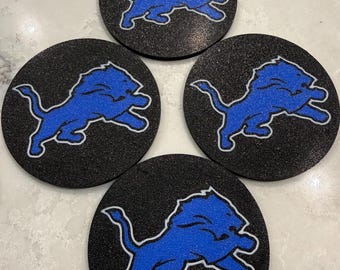 Può includere: Quattro sottobicchieri rotondi neri con superficie strutturata. Ogni sottobicchiere presenta un logo di leone blu con un contorno bianco. Il leone è in posa di corsa. I sottobicchieri sono disposti a quadrato.
