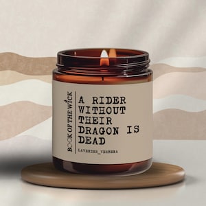 May include: A brown glass candle jar with a white label that reads "Book of the Wick" and "A Rider Without Their Dragon Is Dead" in black text. The candle is lit and the scent is lavender verbena.