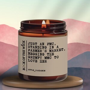 May include: A lit candle in a brown glass jar with a label that reads "JUST AN FMC, STANDING IN A FARMER'S MARKET, BEGGING THE GRUMPY MMC TO LOVE HER." The candle is on a wooden tray. The scent is apple cardamom.