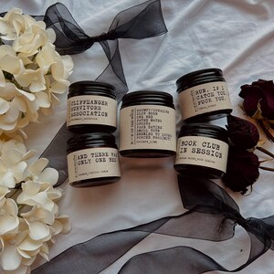 May include: A collection of black-lidded jars with labels, arranged on a white surface with black ribbon and floral accents. The labels feature text such as "Grumpy/Sunshine" and "Book Club in Session". The scene is decorated with white flowers.