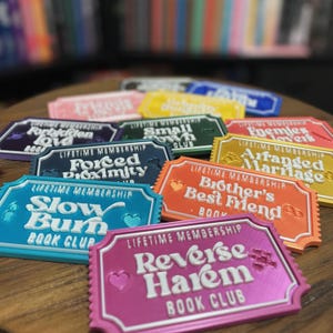 May include: A selection of colourful book club membership badges, each with a unique design and text. The badges feature phrases like "Forbidden Love," "Small Town Club," and "Reverse Harem Book Club." The badges are in various colours, including blue, pink, and orange.