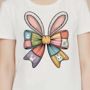 Könnte beinhalten: Weißes T-Shirt mit einem farbenfrohen Oster-Schleifendesign mit Hasenohren. Die Schleife besteht aus Patchwork-Stoff in verschiedenen Farben, darunter Rosa, Grün, Blau und Orange, mit Hasen- und Ei-Motiven.