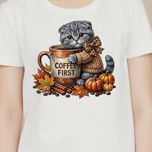 Könnte beinhalten: Ein weißes T-Shirt mit einer Grafik eines grauen Kätzchens, das einen beigen Pullover und eine Schleife mit Leopardenmuster trägt und eine braune Tasse mit dem Text COFFEE FIRST umarmt. Das Motiv zeigt orange Kürbisse, Herbstblätter und Kaffeebohnen.