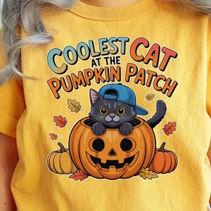 Könnte beinhalten: Senfgelbes T-Shirt mit einer Cartoon-Katze in einer blauen Kappe, die in einem geschnitzten Kürbis sitzt. Der Text lautet "Coolest Cat at the Pumpkin Patch". Enthält Herbstblätter und Kürbisse im Design.
