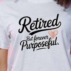 May include: White t-shirt with the words "Retired But forever Purposeful" in black script. A pink heart graphic is between the words "Purposeful" and "But". The shirt is made of a soft material.