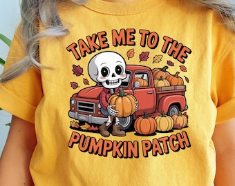 Puede incluir: Camiseta amarilla con un gráfico de un esqueleto sosteniendo una calabaza frente a una camioneta roja llena de calabazas. El texto dice "TAKE ME TO THE PUMPKIN PATCH" en naranja y negro.