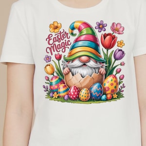 Puede incluir: Camiseta blanca con un diseño de Pascua. Presenta un gnomo saliendo de un huevo roto, rodeado de huevos decorados y flores. El texto "Easter Magic" está encima del gnomo.