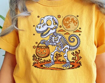 May include: Yellow t-shirt featuring a Halloween-themed graphic. The design includes a white dinosaur skeleton holding a jack-o'-lantern bucket, a full moon, and orange accents. The shirt is made of a soft material.