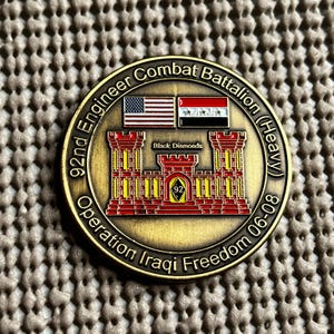 May include: A circular gold-toned challenge coin featuring a red castle emblem with the text 92nd Engineer Combat Battalion (Heavy) and Operation Iraqi Freedom 06-08. It displays the American and Iraqi flags and the text Black Diamonds.