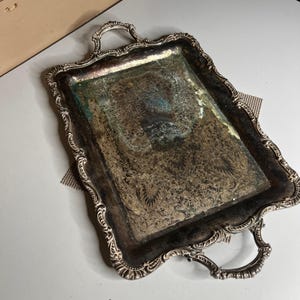 May include: A rectangular silver-toned serving tray featuring ornate, raised scrollwork along the edges and two decorative handles. The central surface displays a detailed, etched floral pattern with visible dark tarnish and oxidation throughout.