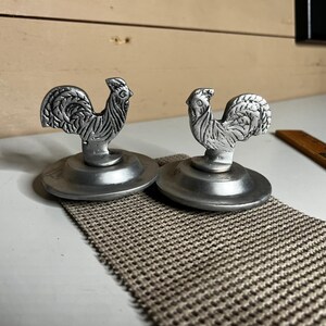 May include: Two silver-colored metal lids, each featuring a sculpted rooster handle. The roosters have detailed feather textures and are mounted on circular, smooth-edged bases. These decorative kitchen accessories are metallic in appearance.