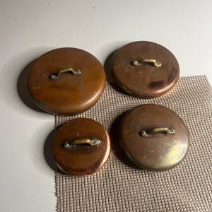May include: Four round copper lids of varying sizes, each featuring a polished brass handle securely attached with two rivets. The copper surfaces exhibit a warm, metallic sheen with natural variations in color and texture across the set.