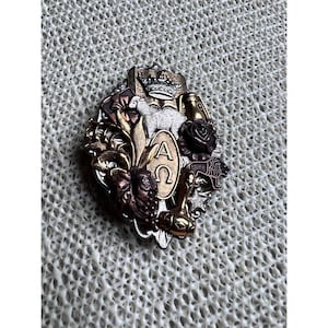 May include: An ornate, multi-toned metal brooch with religious symbols. The brooch features a crown, a dove, a rose, grapes, and the Greek letters Alpha and Omega. The design incorporates gold, silver, and bronze tones, creating a detailed and textured appearance.