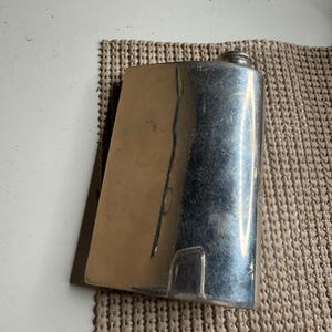 May include: A rectangular silver-toned metal flask with a curved body and a small screw-top cap. The surface of the flask is polished and reflective, showing visible light reflections and minor surface scratches across the metallic exterior.