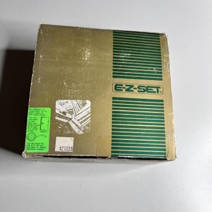 May include: A rectangular cardboard box with the text "E-Z-SET" in green. The box is a light brown color with green stripes and text. There is a sticker on the box with the text "THIS PRODUCT IS ADJUSTABLE TO FIT RIGHT HAND OR LEFT HAND DOORS".