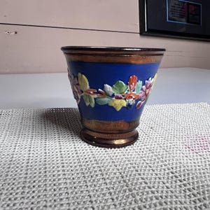 May include: A blue ceramic planter with a floral relief design. The planter has a brown rim and base, with a gold band in the middle. The floral design features colorful leaves and flowers.
