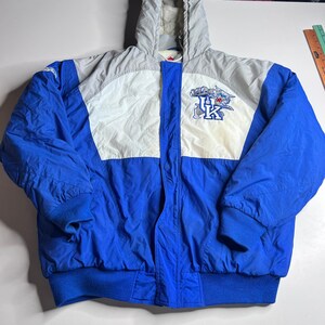 May include: Blue, white, and gray hooded jacket with a full zipper. The jacket features a logo on the chest and blue ribbed cuffs and hem. The jacket is made of a quilted material.