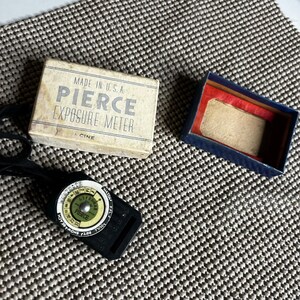 May include: A black Pierce exposure meter with a circular dial featuring white and green markings, accompanied by its original off-white cardboard box labeled MADE IN U.S.A. PIERCE EXPOSURE METER CINE and a separate dark blue empty storage box.
