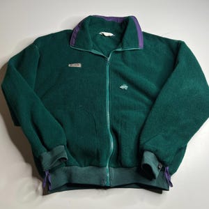 May include: A dark green fleece jacket with a full zipper closure. The jacket features a purple collar and cuffs, with a small logo patch on the chest and a small embroidered design on the pocket. The jacket has two front pockets and a drawstring at the bottom.