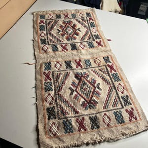 May include: A rectangular woven textile piece featuring two identical square panels with fringed edges. Each panel displays a central diamond motif surrounded by geometric patterns in red, blue, and white thread on a natural beige fabric base.