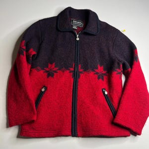 May include: A red and navy blue zip-up jacket with a snowflake pattern. The jacket has a collar, two zippered pockets, and a silver zipper. The brand name "Bernardo Essentials" is visible on a tag inside the collar.