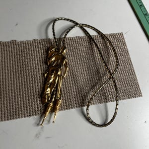 May include: A gold-toned bolo tie featuring a detailed metallic pendant of a figure standing upright with elongated, pointed base elements. The pendant is attached to a thin, braided gold-colored cord that forms a large loop for wear.