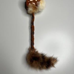 Könnte beinhalten: Ein handgefertigtes Objekt aus braunem Leder und Fell mit einem geflochtenen Griff. Der obere Teil hat ein quadratisches Design mit weißen und braunen Fellakzenten. Der Griff ist mit braunem Leder umwickelt, und der Boden hat einen flauschigen braunen Fellschwanz.