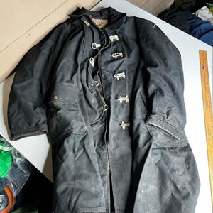 May include: A black long-length coat featuring a front closure with four metal toggles and loops, a high collar, and two large front pockets with flaps. A wooden ruler is placed alongside the coat, showing a length of approximately 36 inches.