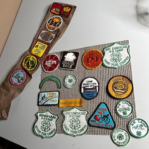 May include: A collection of vintage embroidered fabric patches featuring various shapes, colors, and designs, including 4-H camp emblems, Cookie Olympics badges, and text-based patches, arranged on a tan sash and a textured square mat.