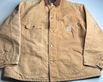 Puede incluir: Chaqueta de trabajo color canela con cuello de pana marrón. La chaqueta tiene cuatro bolsillos delanteros, botones color latón y un parche con el logo de Carhartt en el bolsillo derecho. La chaqueta parece desgastada.