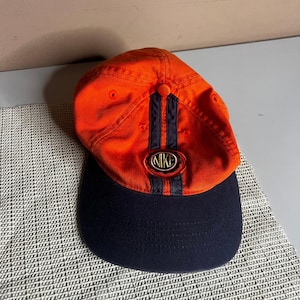 May include: An orange baseball cap with a dark navy blue brim and two vertical navy blue stripes running down the center of the crown. A circular patch featuring the word NIKE in gold lettering is centered on the front of the cap.