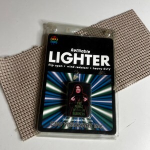 May include: A refillable flip-open wind-resistant lighter in clear plastic packaging. The packaging features the text LIGHTER, C&D Visionary Inc., and an image of a person with the text THE PRINCE OF F***ING DARKNESS in green lettering.