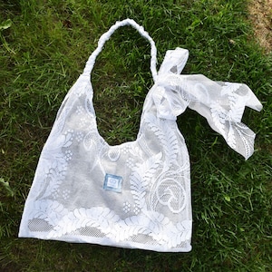 May include: White lace tote bag with a single shoulder strap and a decorative bow. The bag features a floral pattern and a small, square, blue tag. The bag is set against a backdrop of green grass.