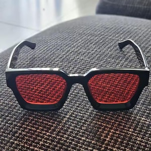 Puede incluir: Gafas de sol negras con lentes rojas. Las gafas tienen una forma geom&eacute;trica y un dise&ntilde;o moderno. Las lentes son de color rojo liso y las monturas son negras. Las gafas de sol son probablemente para moda o estilo.