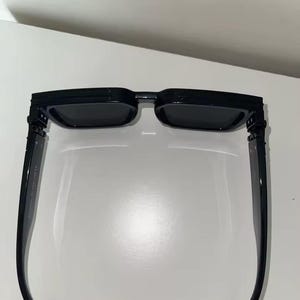 Puede incluir: Gafas de sol rectangulares negras con lentes oscuras. Las gafas tienen una montura negra gruesa y patillas rectas. Las lentes son de un color oscuro s&oacute;lido, y el dise&ntilde;o general es moderno y elegante.