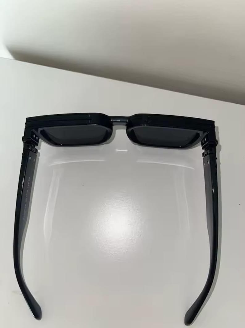Puede incluir: Gafas de sol rectangulares negras con lentes oscuras. Las gafas tienen una montura negra gruesa y patillas rectas. Las lentes son de un color oscuro s&oacute;lido, y el dise&ntilde;o general es moderno y elegante.