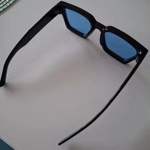 Puede incluir: Gafas de sol negras con lentes azules. Las gafas tienen una montura rectangular y un dise&ntilde;o elegante y moderno. Las lentes son de color azul vibrante y las monturas son de color negro brillante.