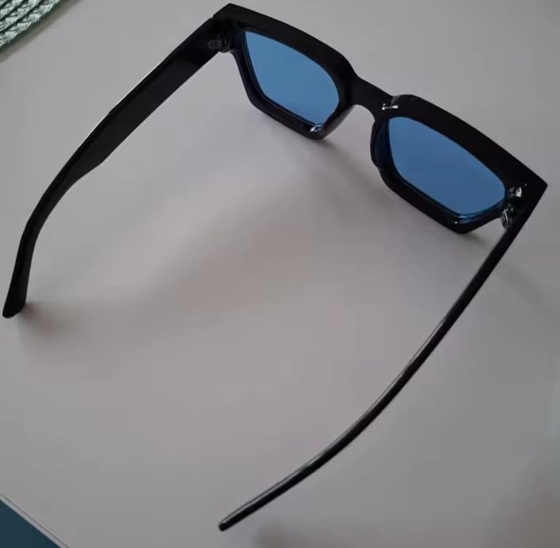 Puede incluir: Gafas de sol negras con lentes azules. Las gafas tienen una montura rectangular y un dise&ntilde;o elegante y moderno. Las lentes son de color azul vibrante y las monturas son de color negro brillante.