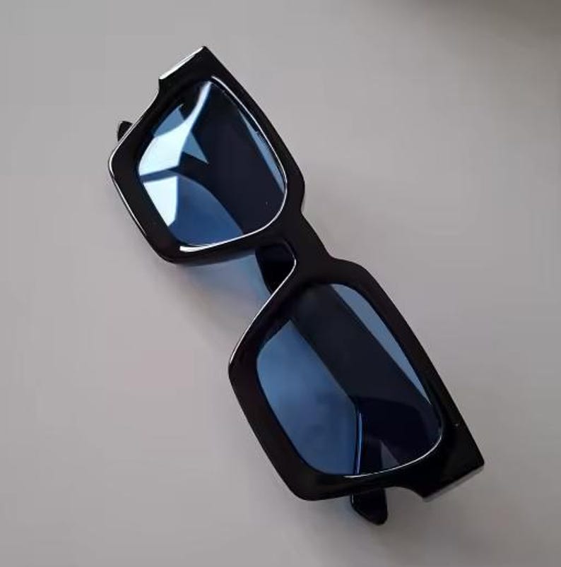 Puede incluir: Gafas de sol negras con una montura cuadrada gruesa y lentes tintadas en azul. Estas gafas de sol tienen un dise&ntilde;o moderno y son adecuadas para todos. Las lentes reflejan la luz, lo que aumenta su apariencia elegante.