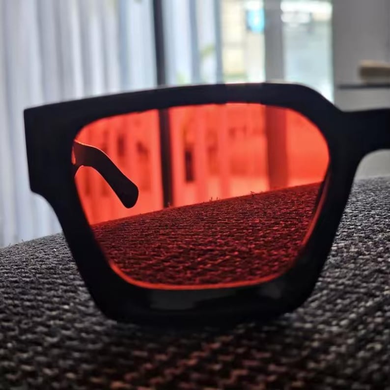 Puede incluir: Gafas de sol cuadradas negras con lentes rojas brillantes. Las gafas tienen una montura gruesa y angular y un dise&ntilde;o elegante y moderno. Las lentes rojas contrastan con la montura negra.