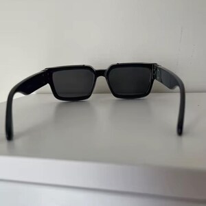 Puede incluir: Gafas de sol negras con montura cuadrada. Las gafas tienen lentes oscuras y patillas negras gruesas. Las gafas est&aacute;n sobre un fondo blanco.