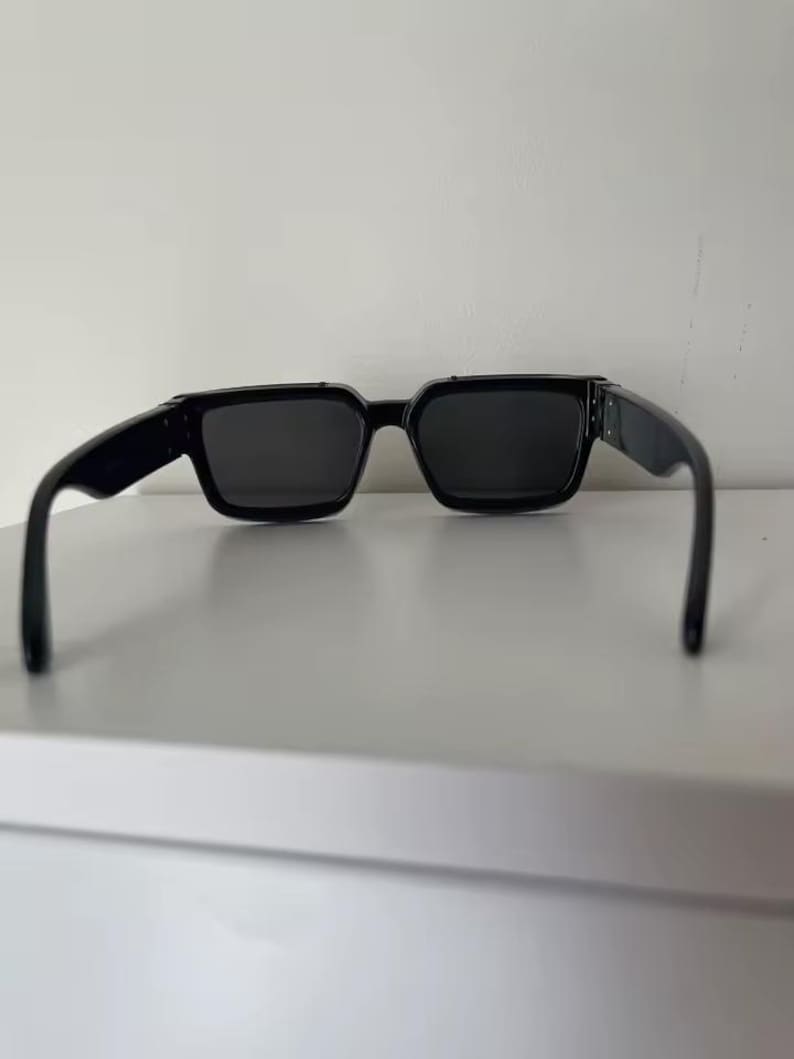 Puede incluir: Gafas de sol negras con montura cuadrada. Las gafas tienen lentes oscuras y patillas negras gruesas. Las gafas est&aacute;n sobre un fondo blanco.