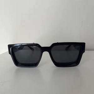 Puede incluir: Gafas de sol negras con montura cuadrada. Las gafas tienen lentes oscuras y una montura gruesa y s&oacute;lida. Las gafas est&aacute;n sobre un fondo blanco liso.