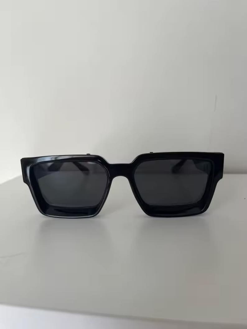 Puede incluir: Gafas de sol negras con montura cuadrada. Las gafas tienen lentes oscuras y una montura gruesa y s&oacute;lida. Las gafas est&aacute;n sobre un fondo blanco liso.