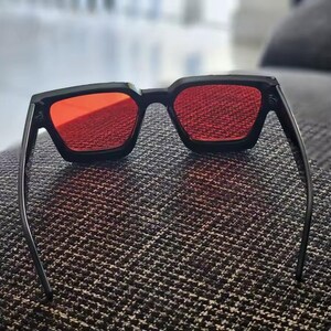 Puede incluir: Gafas de sol negras con lentes rojas. Las gafas tienen una forma cuadrada y un dise&ntilde;o elegante y moderno. Las lentes son de color rojo vibrante, ofreciendo un contraste audaz con la montura negra. Las patillas son delgadas.