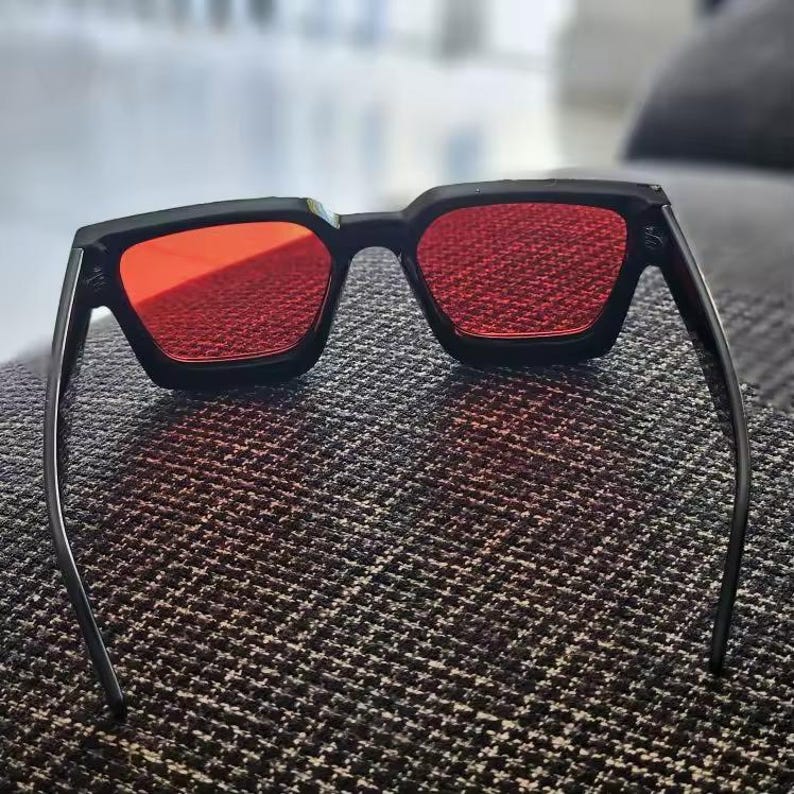 Puede incluir: Gafas de sol negras con lentes rojas. Las gafas tienen una forma cuadrada y un dise&ntilde;o elegante y moderno. Las lentes son de color rojo vibrante, ofreciendo un contraste audaz con la montura negra. Las patillas son delgadas.