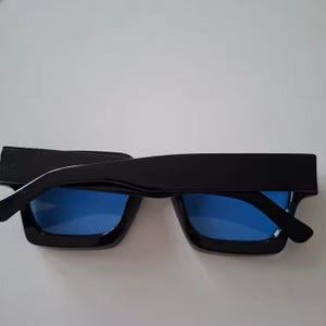 Puede incluir: Gafas de sol negras con montura gruesa y lentes cuadradas. Las lentes son de color azul brillante. Las gafas de sol se ven desde arriba, mostrando el dise&ntilde;o de la montura.